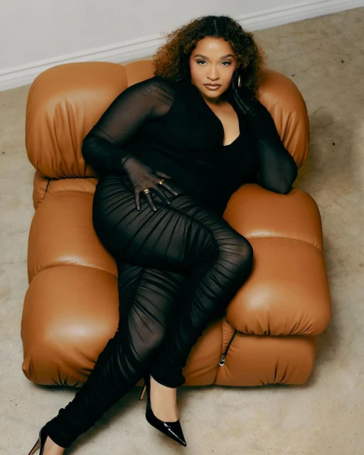 Woman in a black catsuit poses on a tan leather lounge chair, wearing black high heels and gold jewelry, against a neutral background.