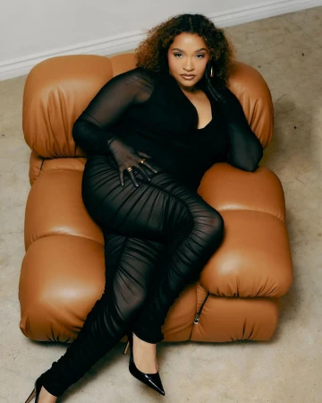 Woman in a black catsuit poses on a tan leather lounge chair, wearing black high heels and gold jewelry, against a neutral background.