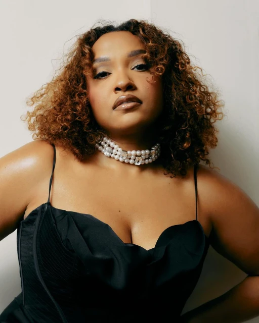 Portrait of an adult woman with curly red hair wearing a black dress and pearl choker necklace against a neutral studio backdrop.