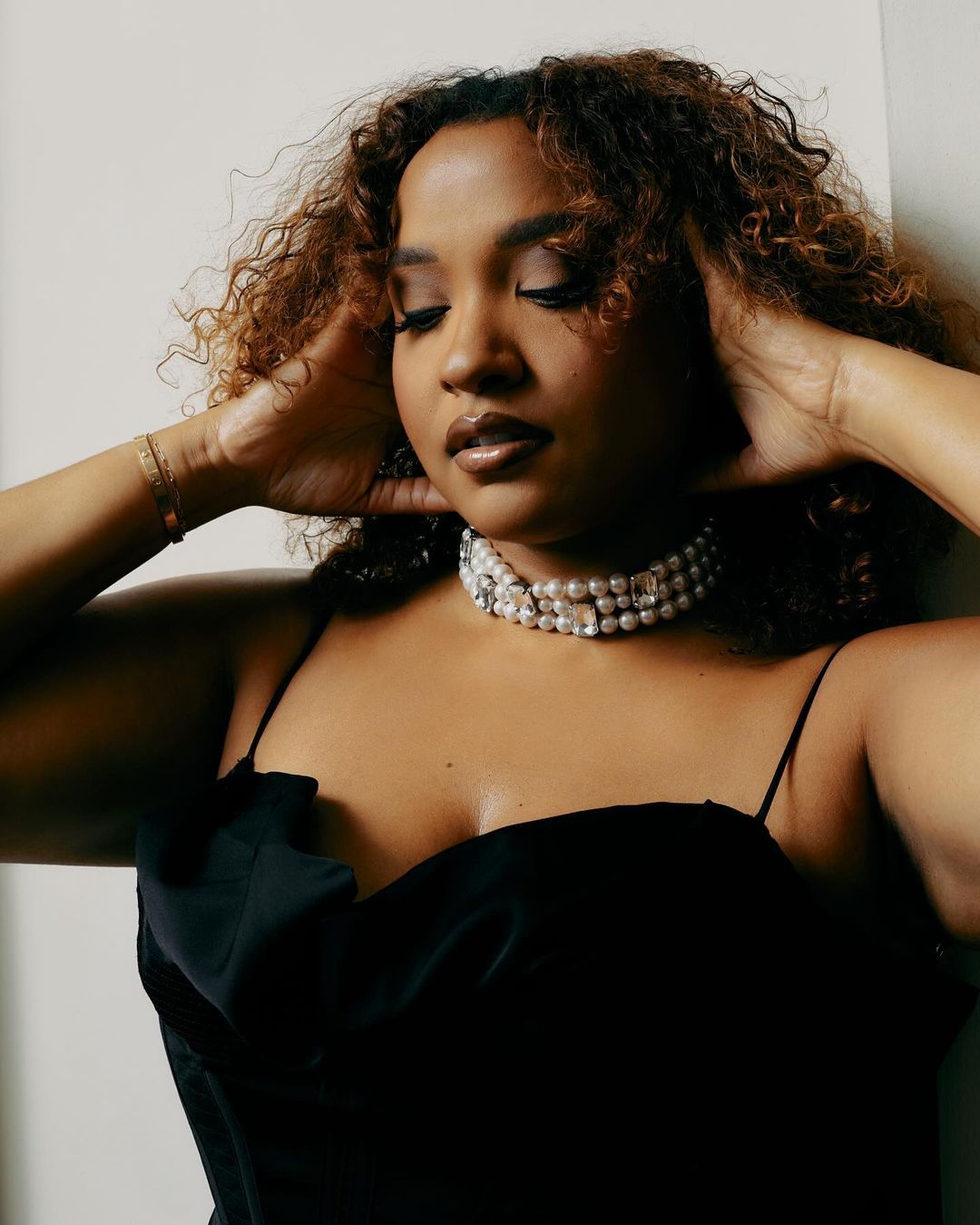 Elegant portrait of a young woman with curly hair wearing a black top and pearl choker, posing against a light background.