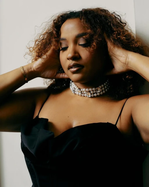 Elegant portrait of a young woman with curly hair wearing a black top and pearl choker, posing against a light background.