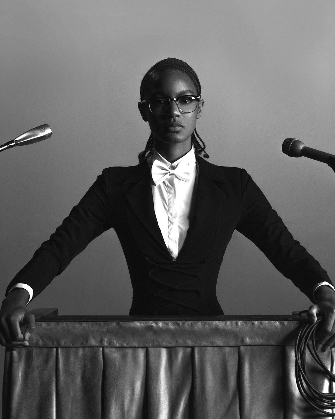 Glamorous portrait of a Black woman in a stylish black blazer and white bow tie, standing at a podium with microphones in a studio.