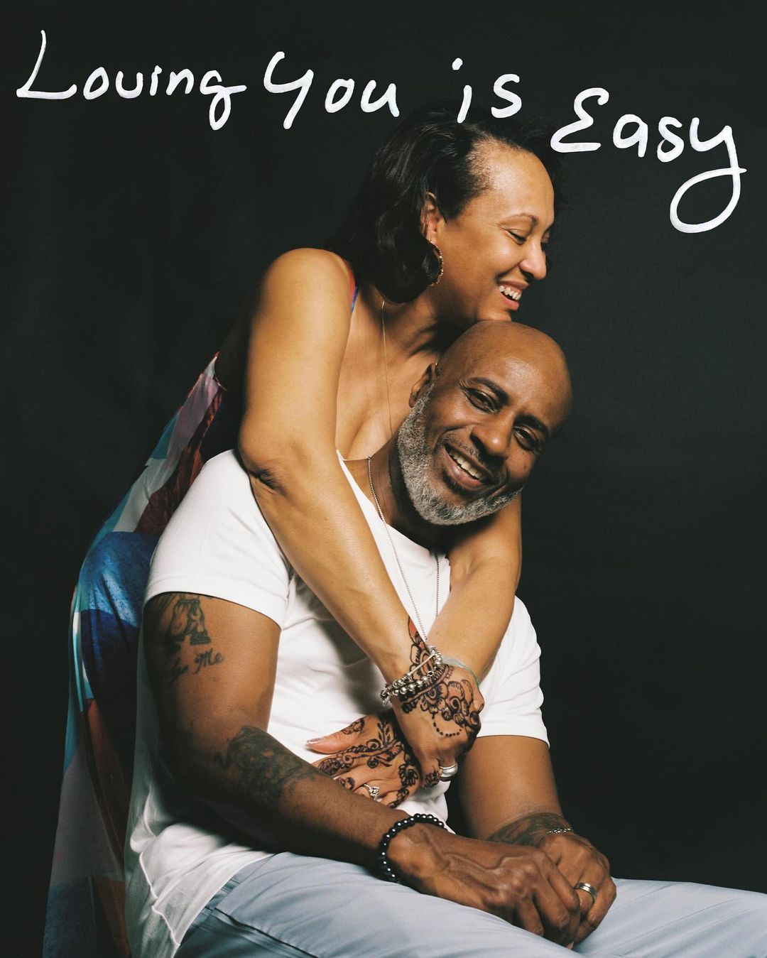 Happy couple embraces in a close-up shot; the woman wraps her arms around the man, both smiling against a dark background, studio lighting.