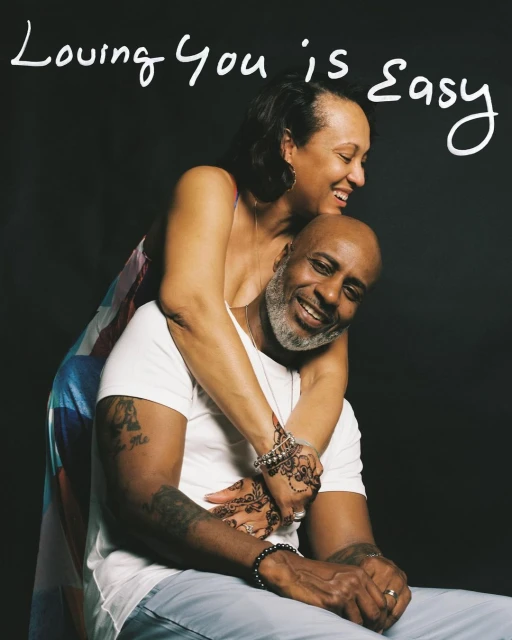 Happy couple embraces in a close-up shot; the woman wraps her arms around the man, both smiling against a dark background, studio lighting.