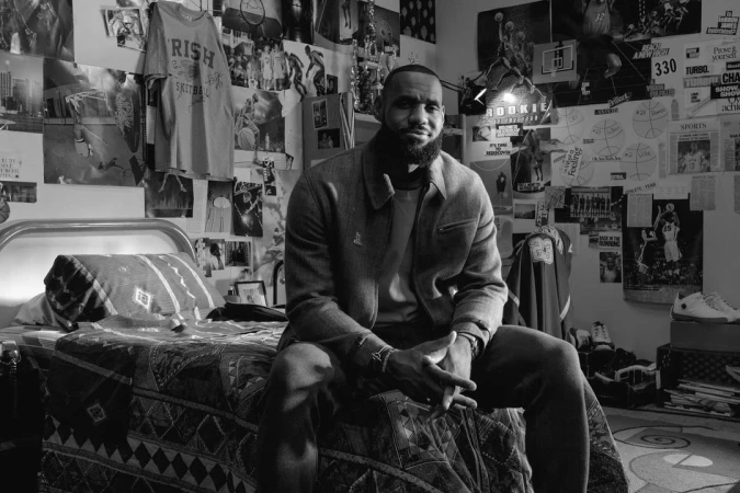Monochrome portrait of a bearded man sitting on a patterned bed in a room with basketball-themed posters and memorabilia on the walls.