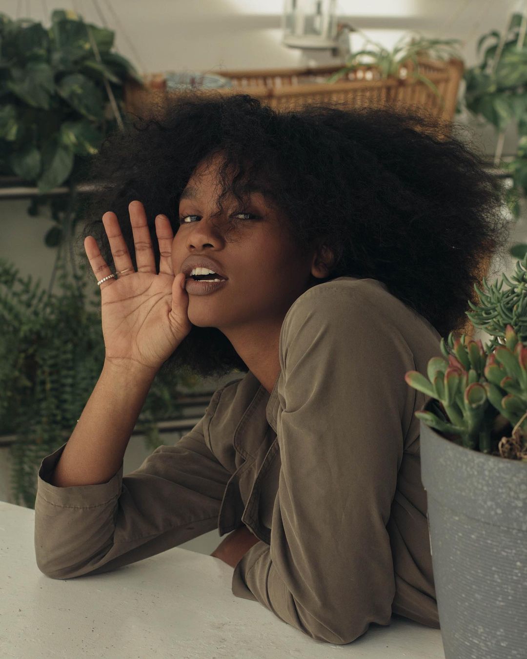 Portrait of an African American woman with an afro calling out, wearing a khaki shirt, in a bright indoor setting with plants.
