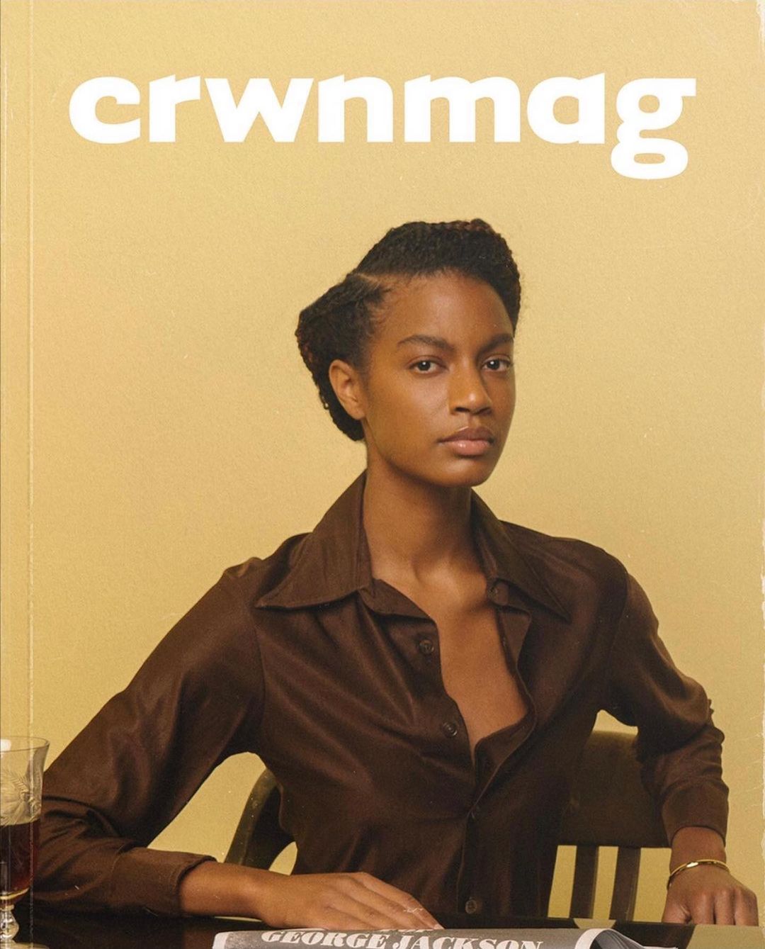 Magazine cover featuring a Black woman with dark hair, wearing a brown collared shirt, at a table with a drink and newspaper.