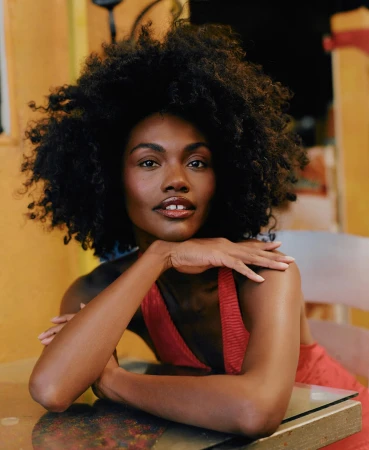 Portrait of a smiling Black woman with voluminous curly hair wearing a red top, resting her chin on her hands.