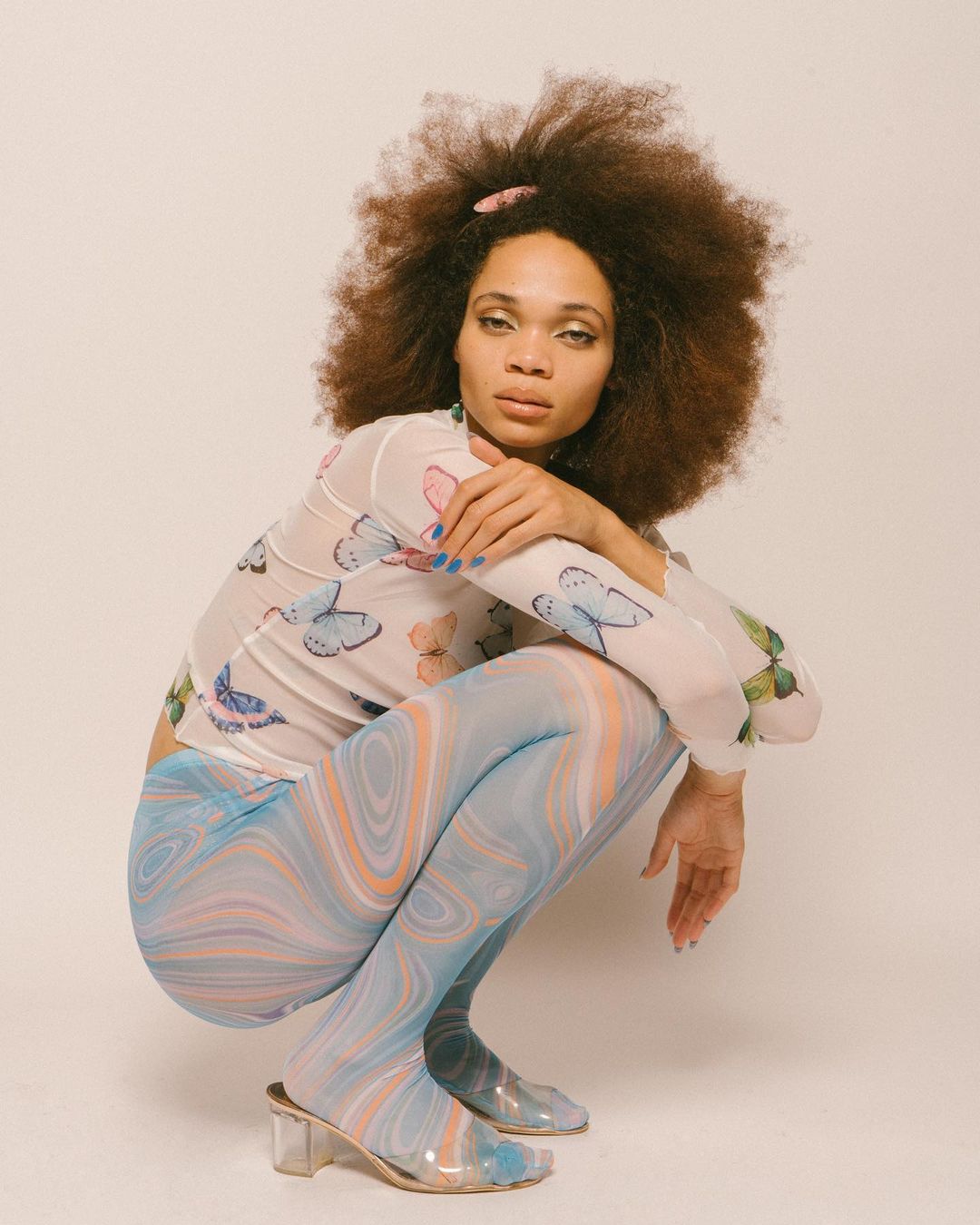 A young adult woman with afro-textured hair and butterfly patterned top poses in a photo studio while wearing marble pattern tights.