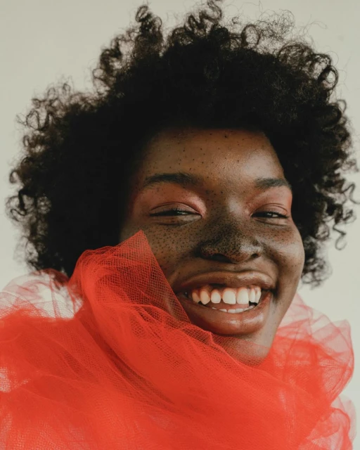 Smiling Black woman with curly hair, wearing a red tulle scarf, showcases vibrant eye makeup in a studio close-up portrait on a light backdrop.