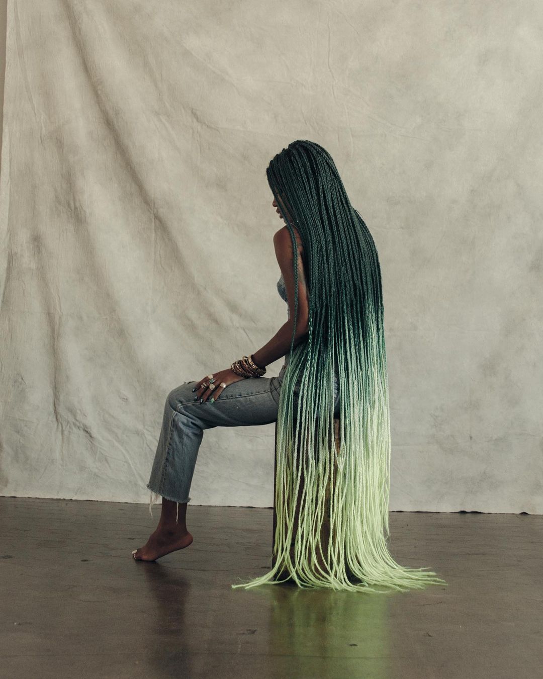 Young woman with long green-to-blonde ombre box braids sitting on a stool in front of a neutral backdrop in a studio environment.