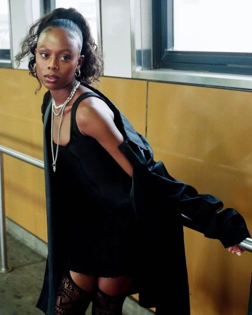 Young adult woman in a black dress and coat poses on stairs in an urban setting, wearing jewelry and hosiery.