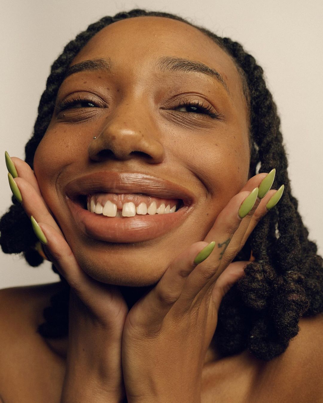 Close-up of a smiling African-American woman with dreadlocks and green nails, hands framing her face, against a neutral background.