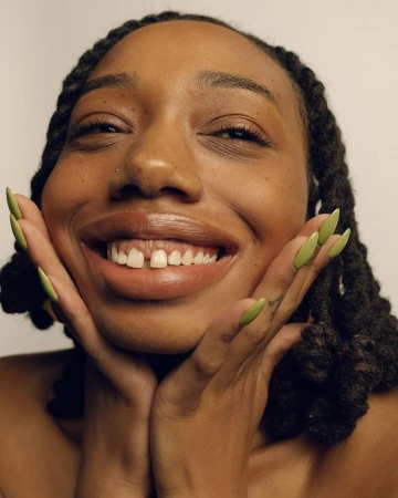 Close-up of a smiling African-American woman with dreadlocks and green nails, hands framing her face, against a neutral background.
