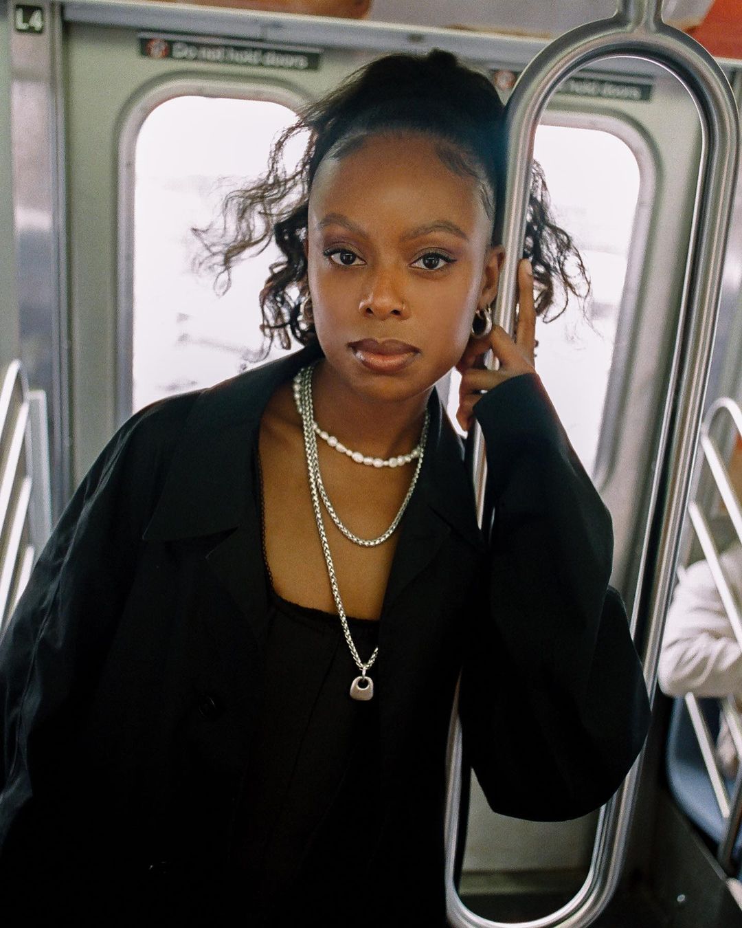 A young Black woman in a black jacket and jewelry stands on a subway train, holding onto the rail, with a neutral expression on her face.
