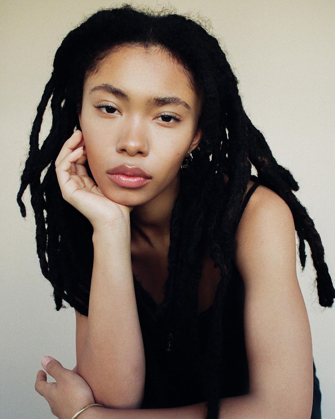 Portrait of a young woman with long black dreadlocks resting her chin on her hand against a neutral background.