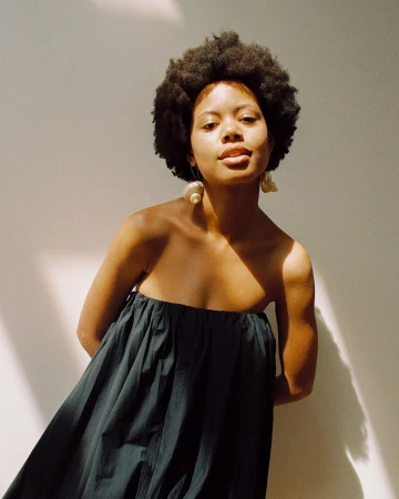 Portrait of a woman with natural hair in a dark strapless dress against a white wall, illuminated by natural sunlight, featuring statement earrings.