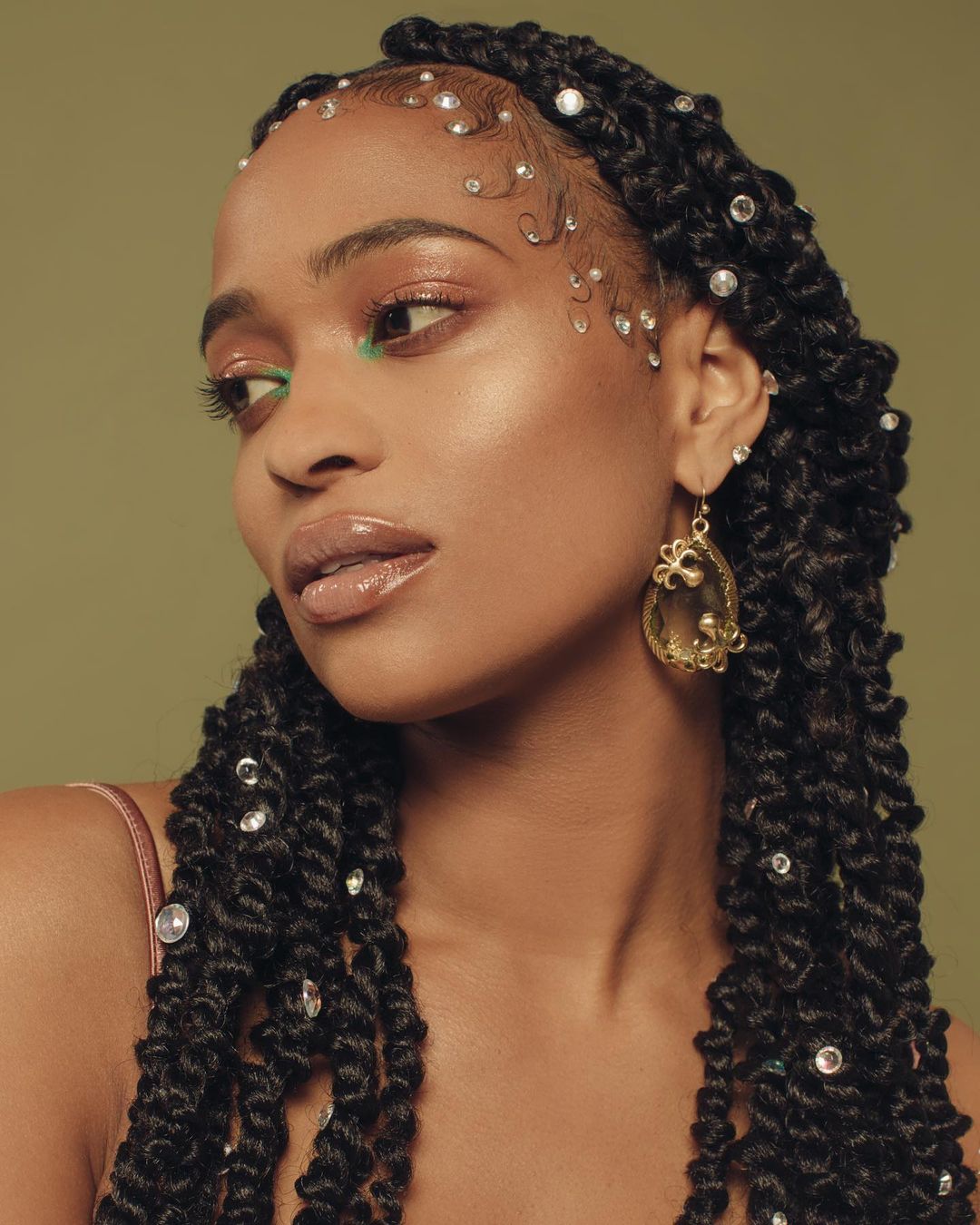Portrait of a woman with decorated passion twists, makeup, and gold earrings against a soft olive green backdrop in a beauty editorial shot.
