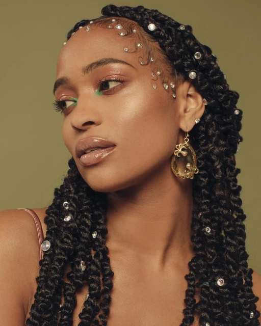 Portrait of a woman with decorated passion twists, makeup, and gold earrings against a soft olive green backdrop in a beauty editorial shot.