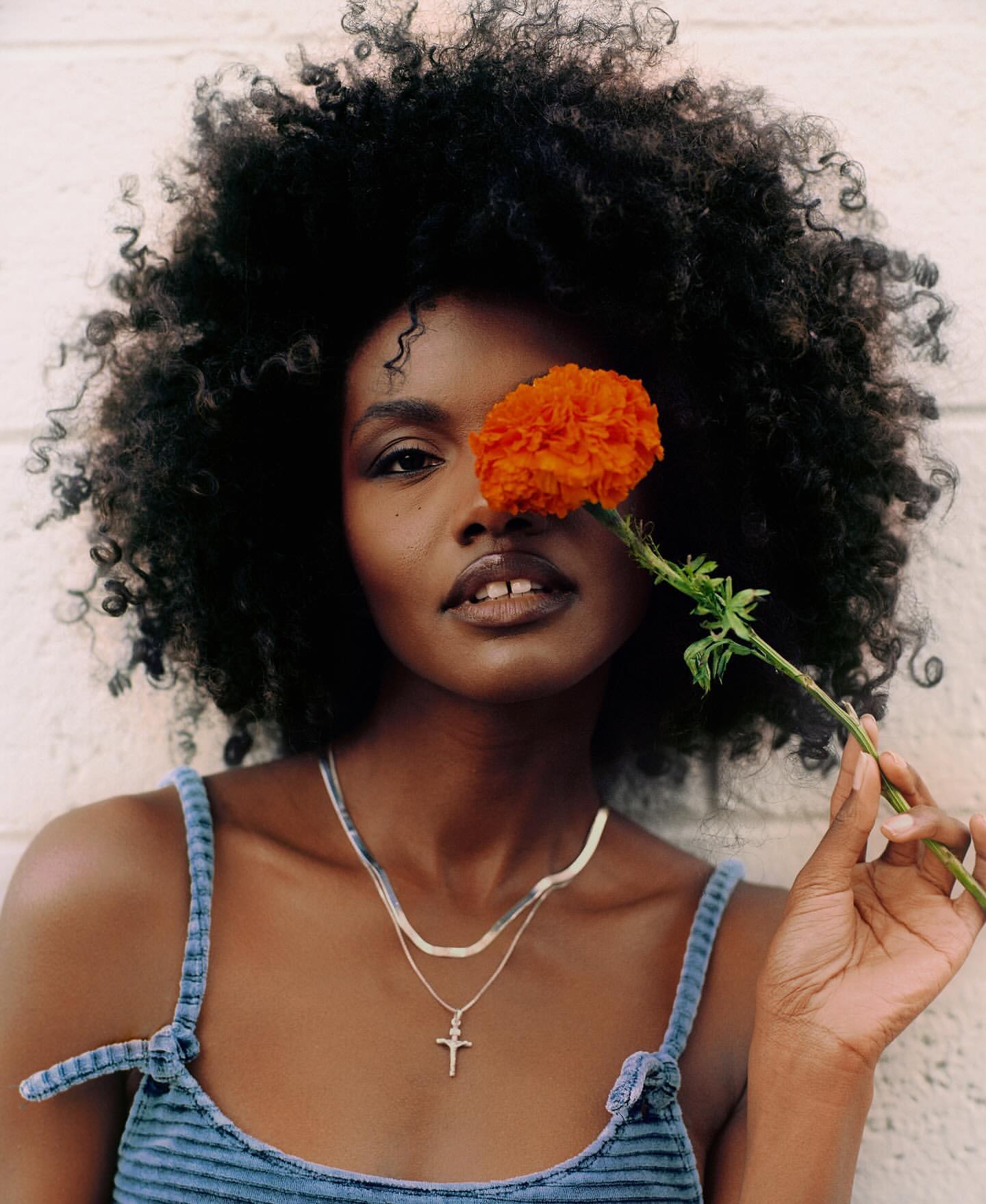Portrait of a Black woman holding a vibrant orange flower, showcasing her dark hair and silver necklaces against a white wall.