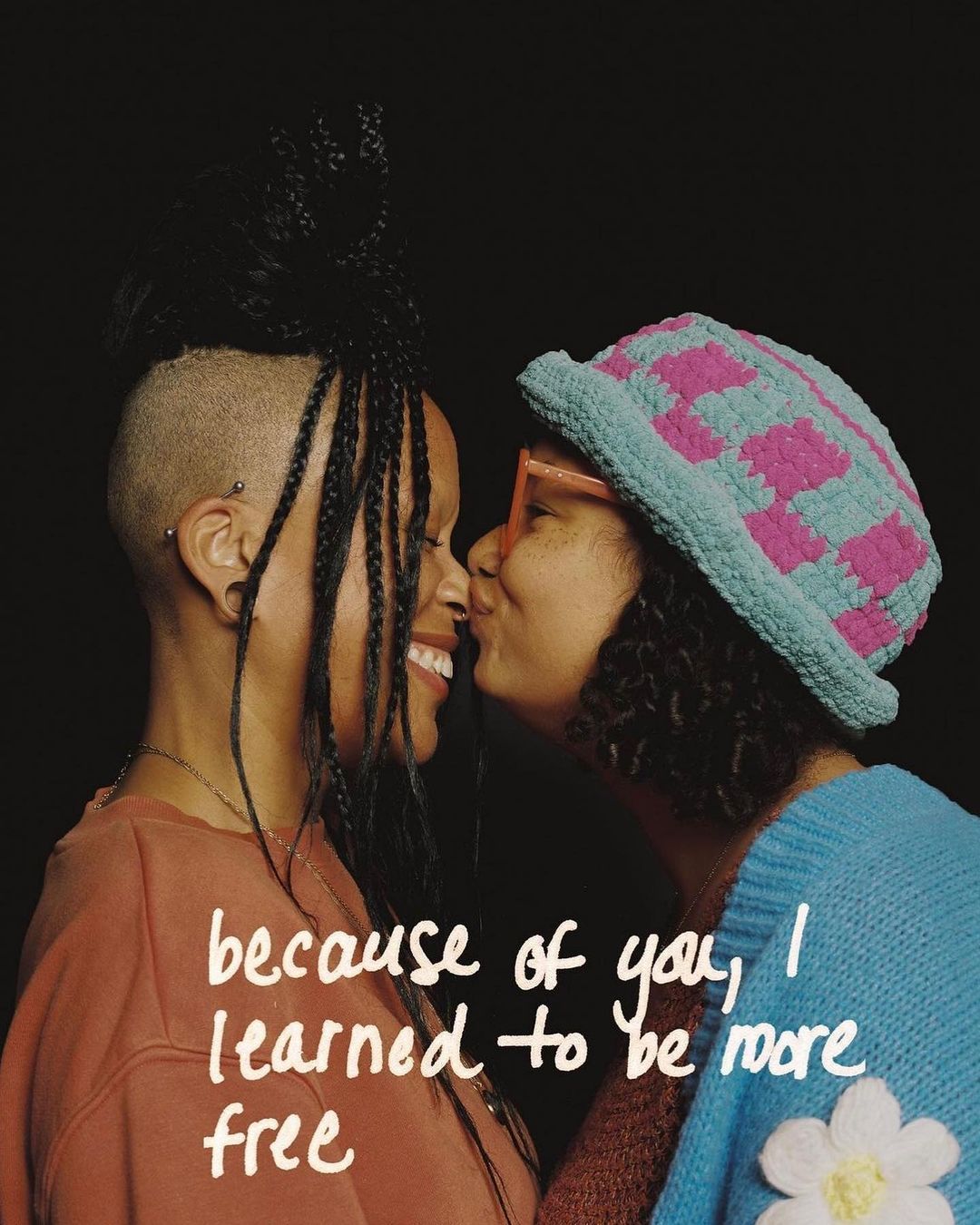 Two women with braids, one with a crocheted hat, stand closely and touch noses in a tender moment against a dark background with text.