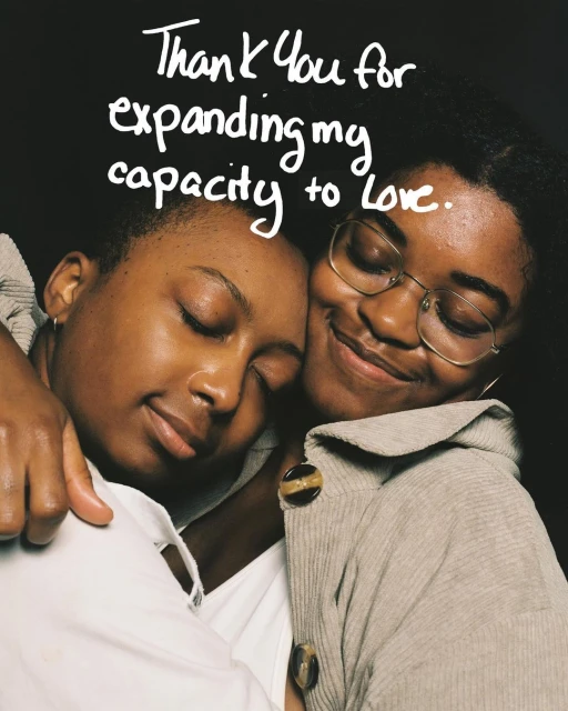 Two African-descent women embrace warmly in a close-up shot, one wearing glasses and a tan sweater against a dark backdrop.