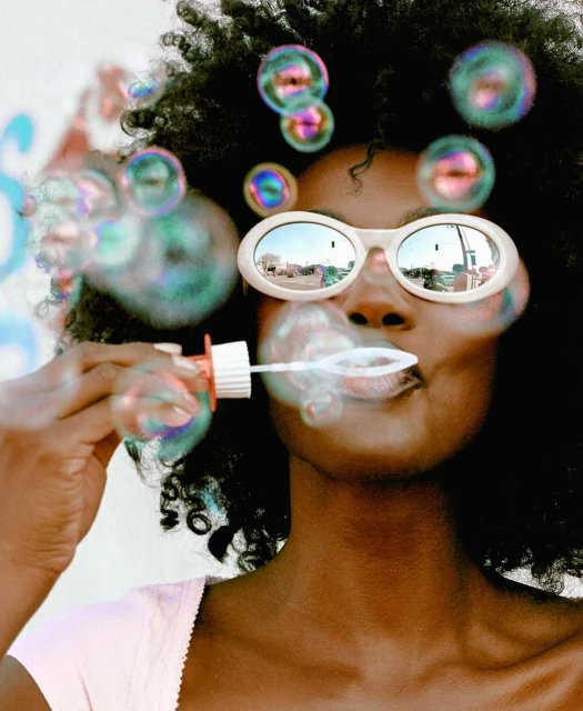 A woman with afro puffs bubbles in sunglasses reflecting a street scene, wearing a pastel top and pale pink lipstick, in front of a wall.