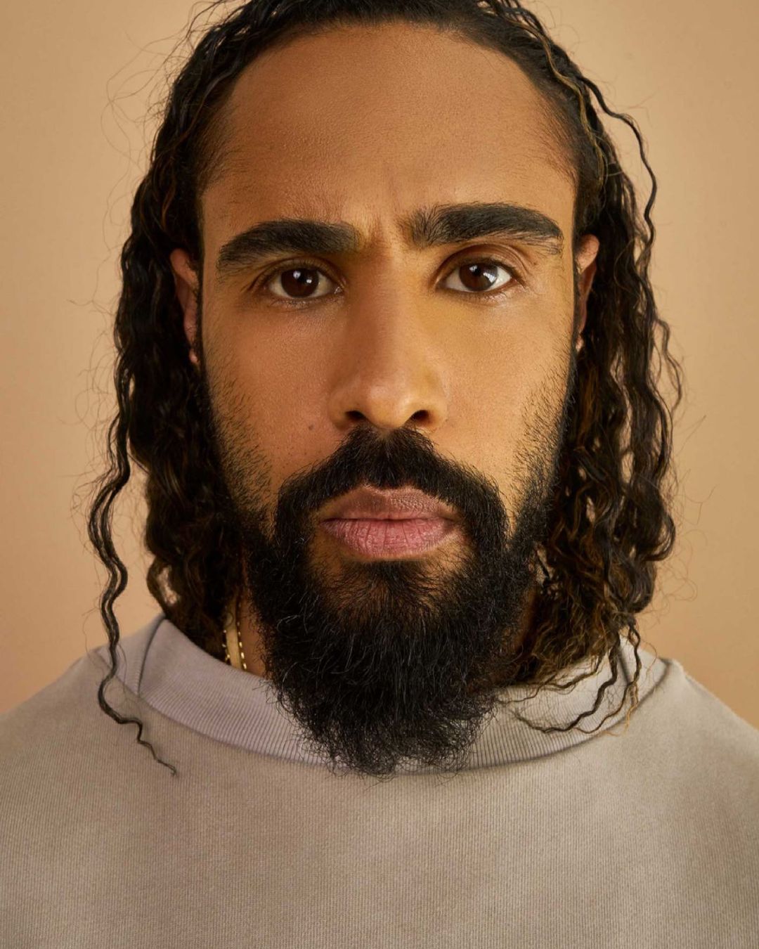 Close-up portrait of an adult male with long, curly hair and a beard, wearing a beige turtleneck sweater against a beige backdrop.