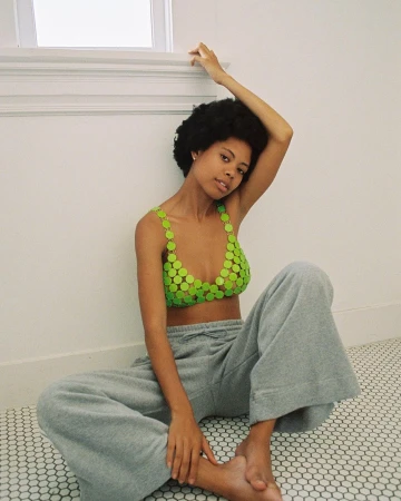A young adult woman with an afro sits on a tiled floor, wearing a green top and gray sweatpants, posing near a window with natural light.
