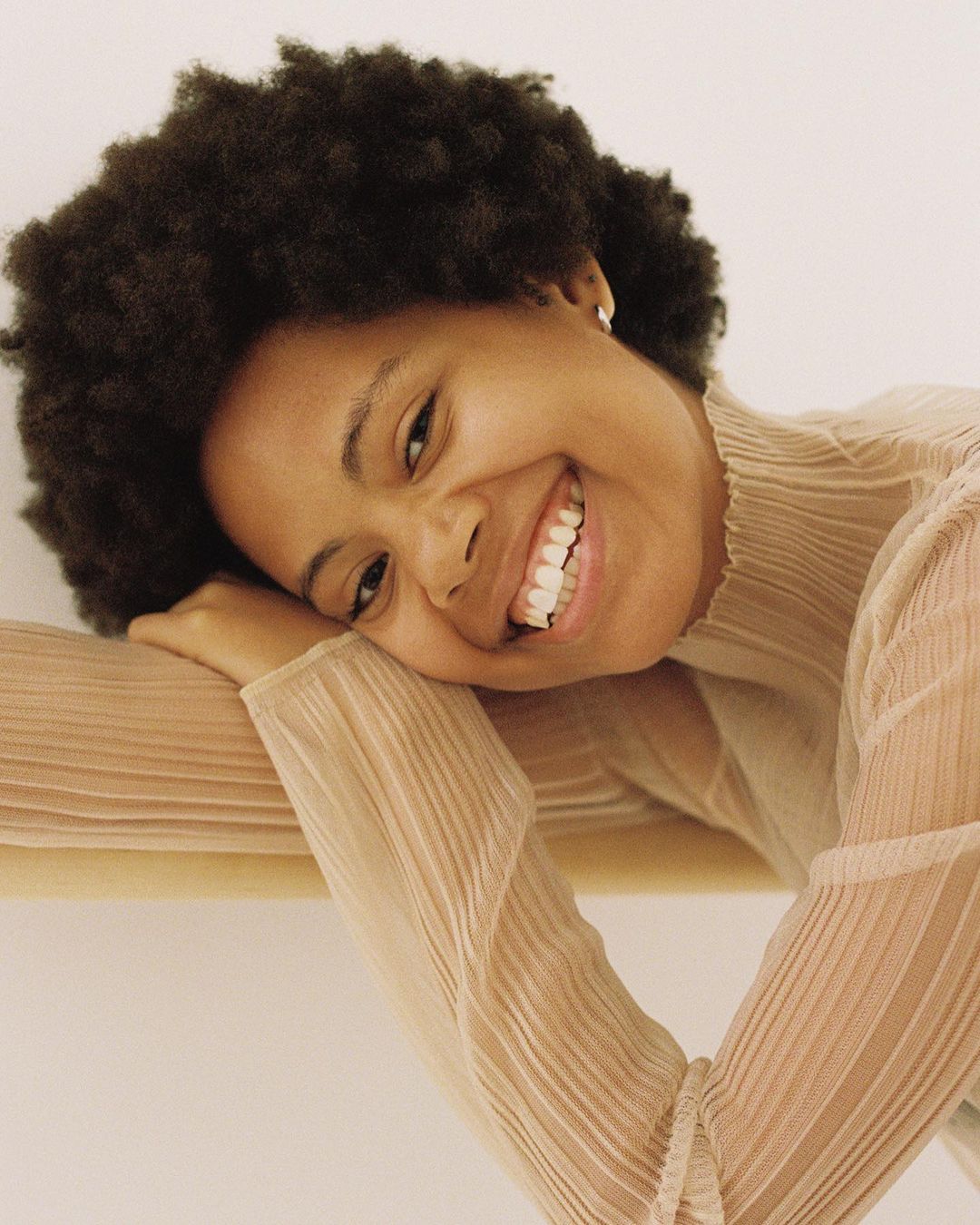 Portrait of a smiling Black woman with curly hair resting her head on her arm, wearing a sheer beige top.