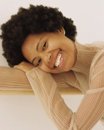 Portrait of a smiling Black woman with curly hair resting her head on her arm, wearing a sheer beige top.