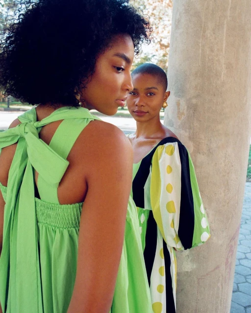 Two black female fashion models in green and polka dot print dresses pose outdoors with soft natural lighting, showcasing summer style.