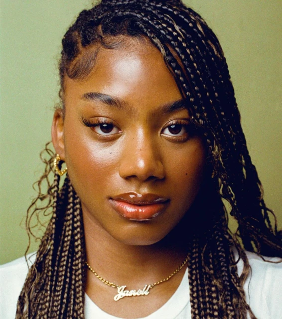Portrait of a dark-skinned young woman with braided hair, gold hoop earrings, and a nameplate necklace against a green backdrop.