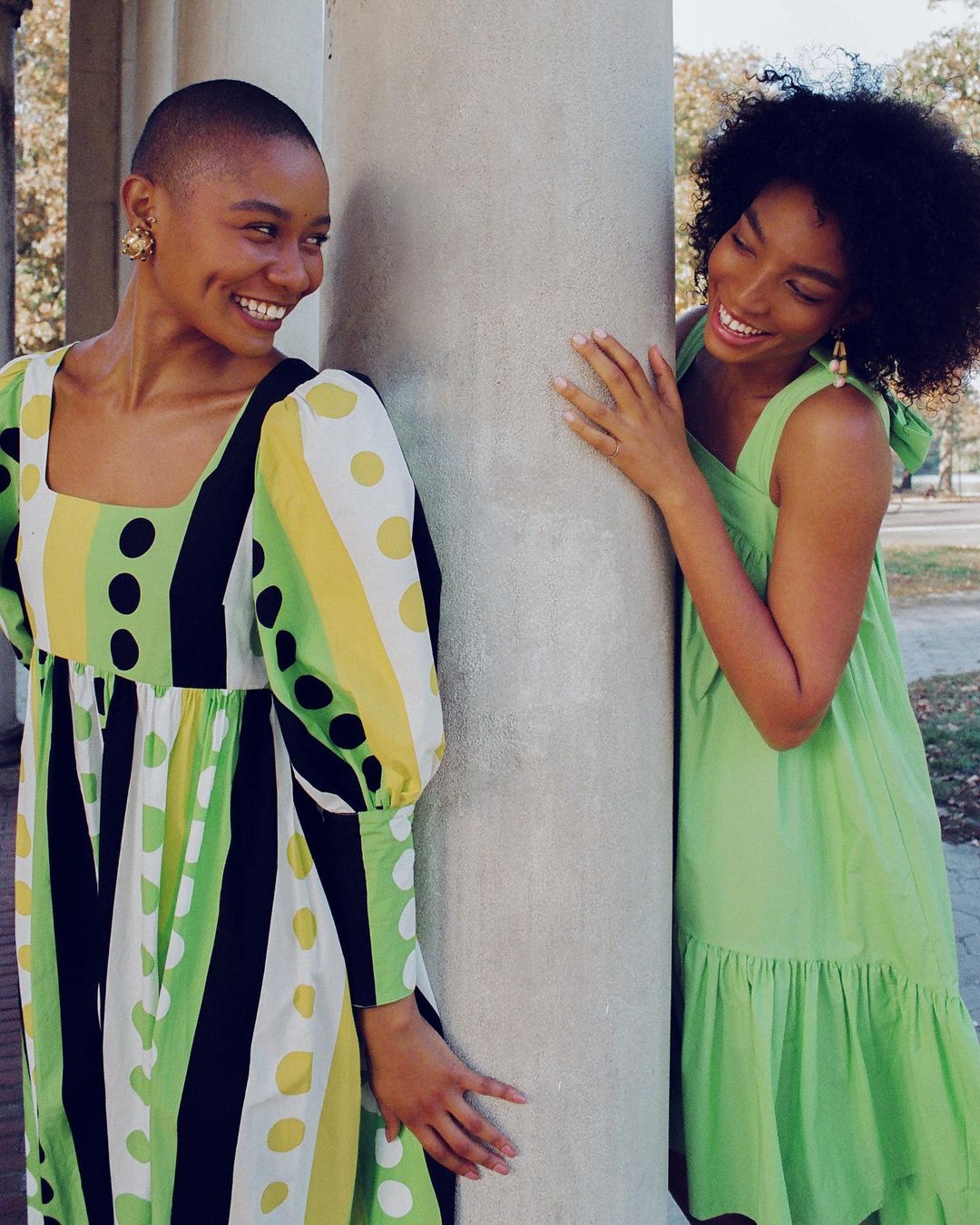 Two smiling African-American women in bright dresses pose near a column outdoors, showcasing fashion and friendship in natural light.