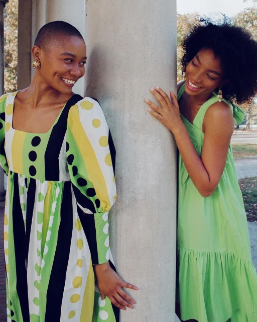Two smiling African-American women in bright dresses pose near a column outdoors, showcasing fashion and friendship in natural light.