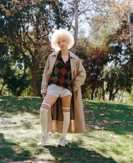 Young woman with an afro poses in a park wearing a trench coat, patterned shirt, shorts, knee socks and sneakers, with trees in the background.
