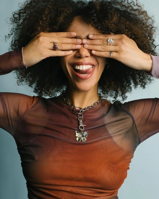 Young woman with curly hair covers her eyes with her hands, sticking her tongue out, wearing a butterfly pendant necklace and rings.