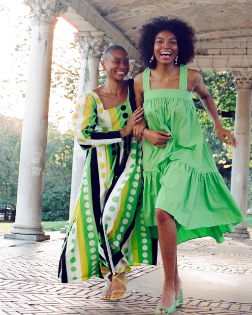Two women in vibrant summer dresses walk on a stone path under a colonnade, smiling brightly with a sunlit green background.
