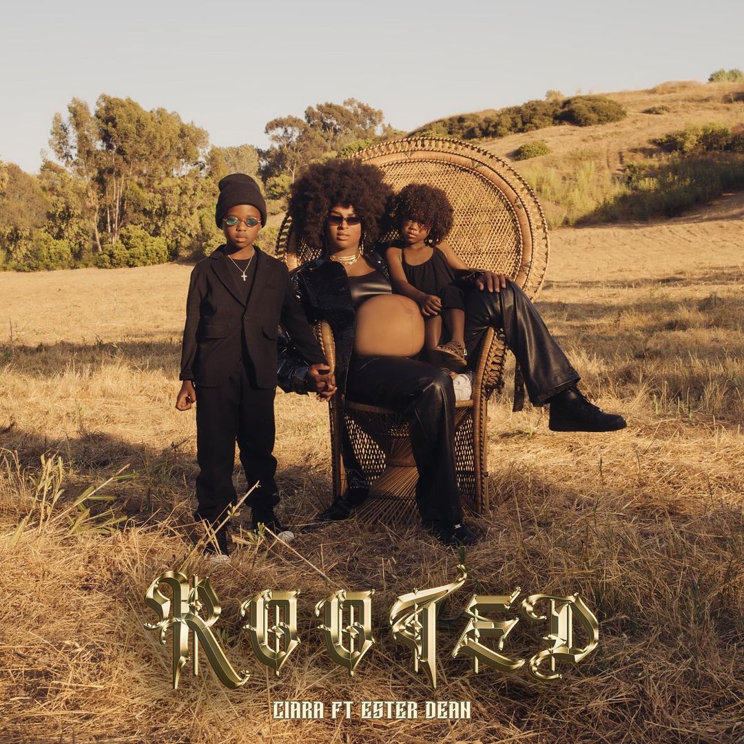 Pregnant woman seated with two children in a field, all wearing black outfits, against a backdrop of trees and a blue sky.