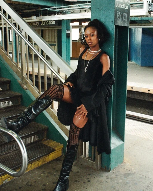 Woman in a black dress and boots poses on subway stairs with a black jacket slung over her shoulder. Necklace and green accents.