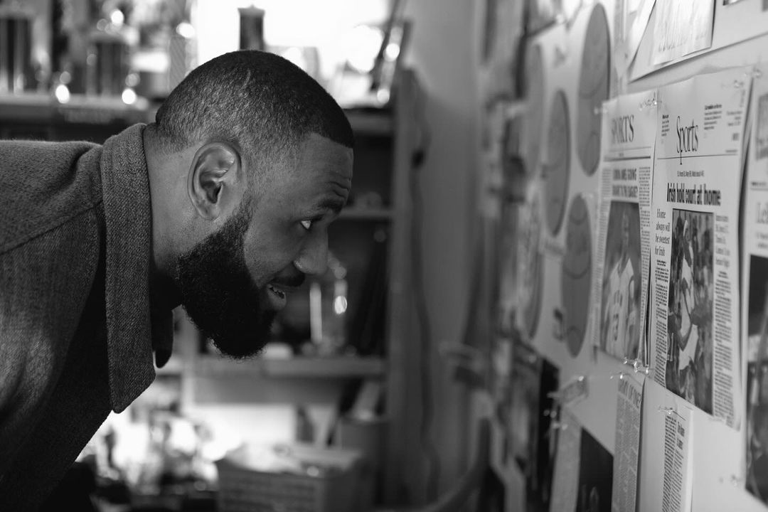 Black and white profile shot of a Black man looking at newspaper clippings on a wall, with shelves and various objects blurred in the background.