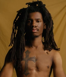 Portrait of a young Black man with long locs and facial and chest tattoos against a mustard yellow backdrop.