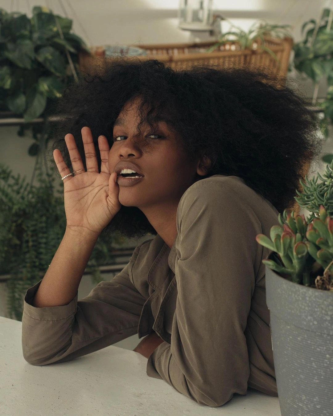 Portrait of a Black woman with voluminous dark curly hair holding her hand to her cheek against a plant-filled indoor background