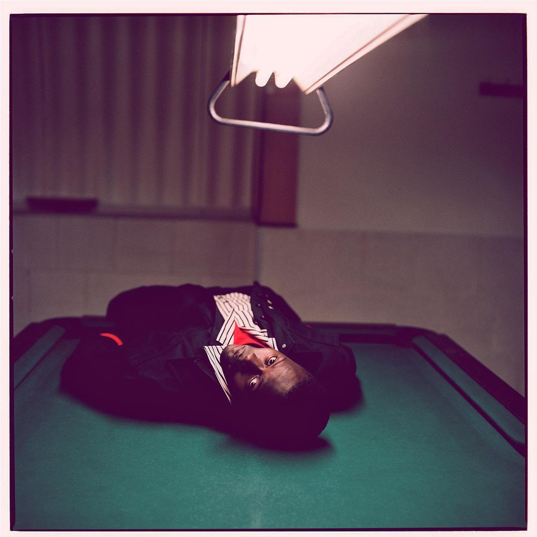 An adult male laying on a green billiard table under a bright overhead light in an indoor setting, wearing a dark jacket and red undershirt.