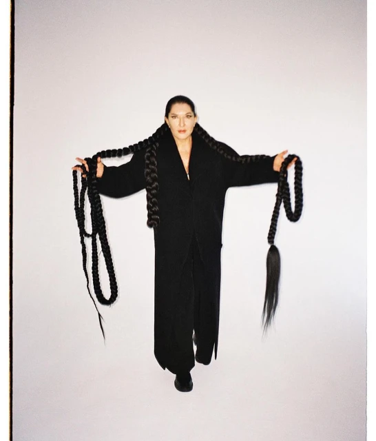 A woman with long braided hair stands against a white background, wearing a long black coat and black pants in a studio shot.