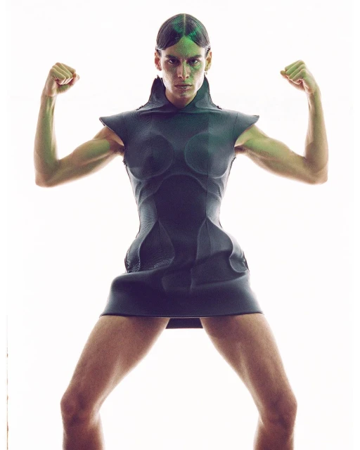 A model with green lighting poses in a short, dark dress, flexing both arms while standing against a bright white backdrop.