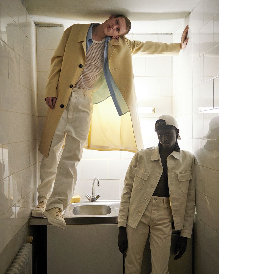 Two male models in a narrow tiled space with a sink, one wearing a yellow coat and white trousers, the other in a cream jacket.