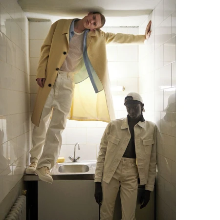 Two male models in a narrow tiled space with a sink, one wearing a yellow coat and white trousers, the other in a cream jacket.
