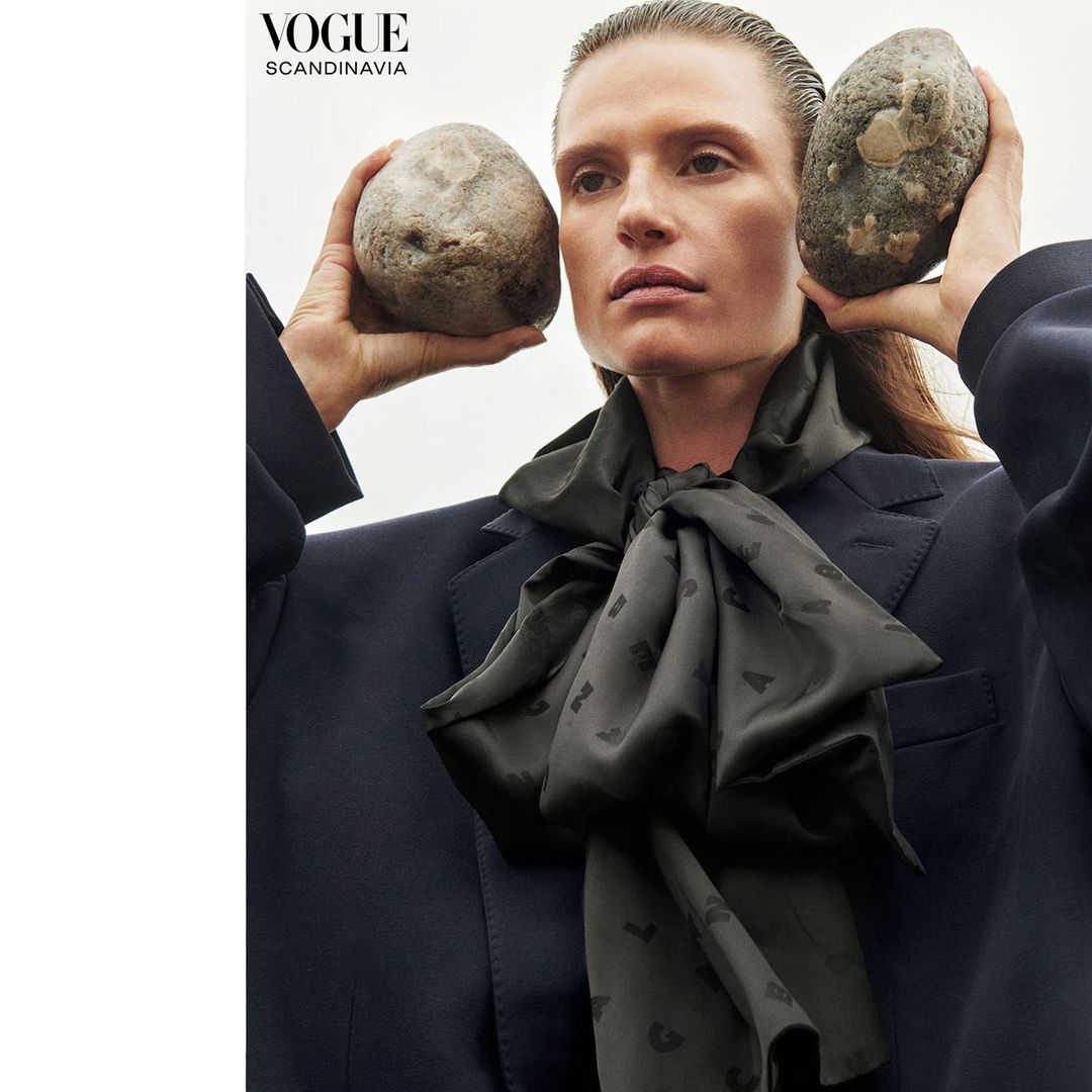 Fashion editorial featuring a woman holding rocks, wearing a black blazer, and a patterned scarf, against a simple white background.