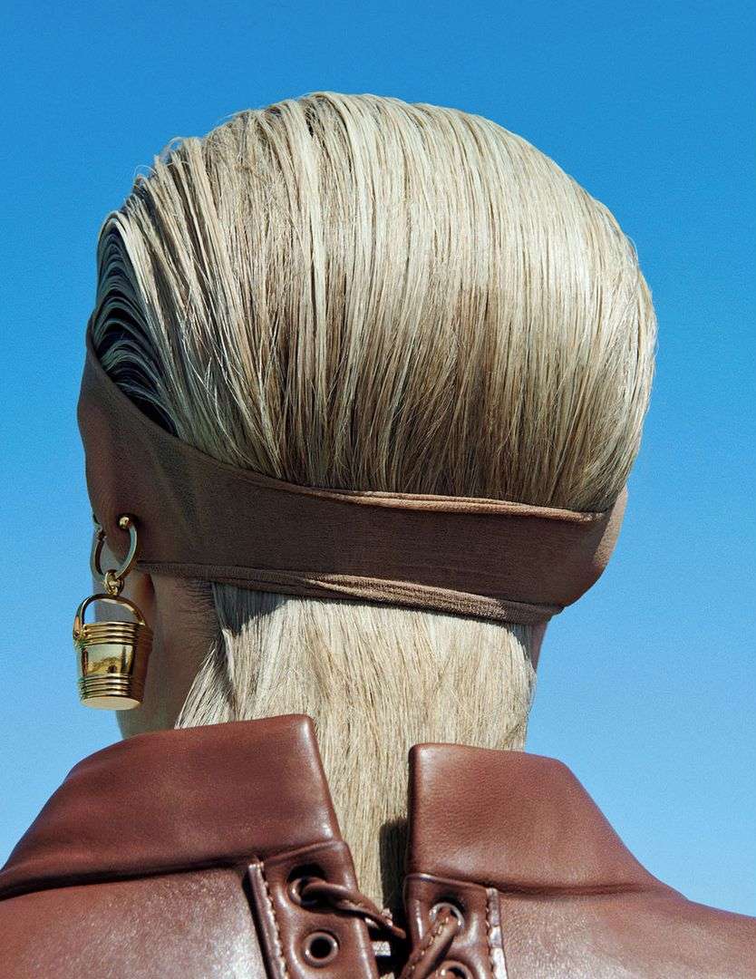Back view of a woman with blonde hair wearing a brown headband and a brown leather jacket against a clear blue sky.